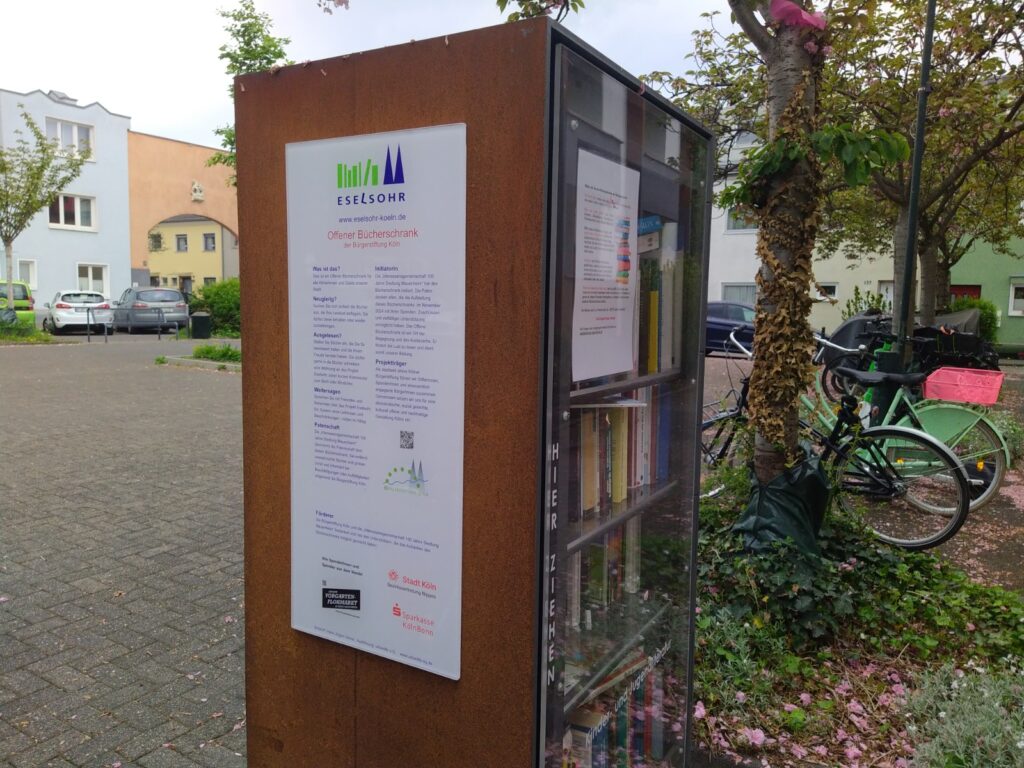 Unser öffentlicher Bücherschrank auf dem Nibelungenplatz von der Seite im Vordergrund mit den Hinweisen auf die Spendenden - im Hintergrund ist der Torbogen am Nibelungenplatz zu sehen.
